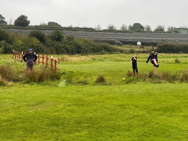 Two Footgolf Devon League players watching another play mid way through kicking the football into the feild