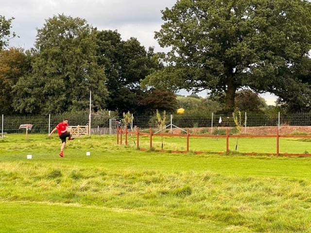 Young man with a red t-shirt and black shorts kicking a ball from far away in a green field